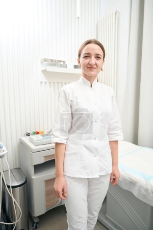 Téléchargez les photos : Pleine longueur vue de face portrait de charmante femme médecin caucasienne est debout dans son cabinet et posant à l'appareil photo - en image libre de droit