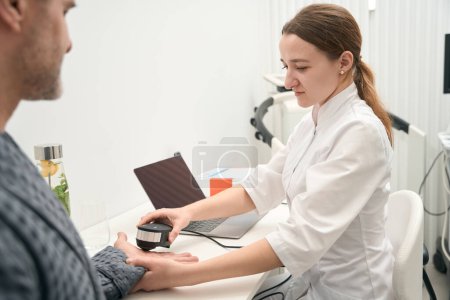 Photo for Waist up side view portrait of focused elegant Caucasian doctor is using oligoscan for examining body indicators in medicine clinic - Royalty Free Image