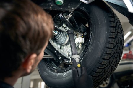 Foto de Hombre en ropa protectora de pie en el montaje de neumáticos debajo del coche y la inspección de los collares de arranque - Imagen libre de derechos