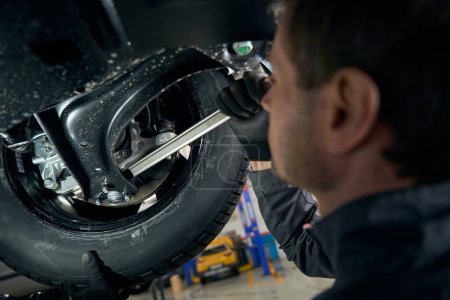 Téléchargez les photos : Réparateur en vêtements de protection debout dans le montage des pneus sous la voiture et l'inspection des colliers de démarrage - en image libre de droit