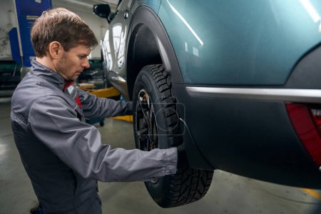 Foto de Mecánico en ropa protectora de pie en el montaje de neumáticos cerca del coche y comprobación de rodamientos de ruedas escorrentía - Imagen libre de derechos