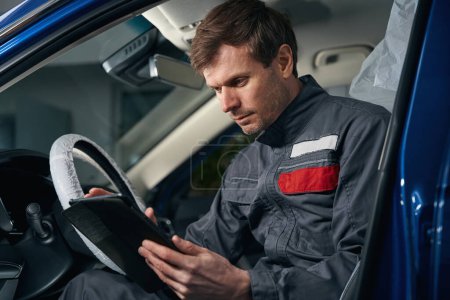 Téléchargez les photos : Homme en vêtements de protection assis dans la voiture, tenant l'ordinateur portable et éliminant les erreurs du système informatique de bord - en image libre de droit