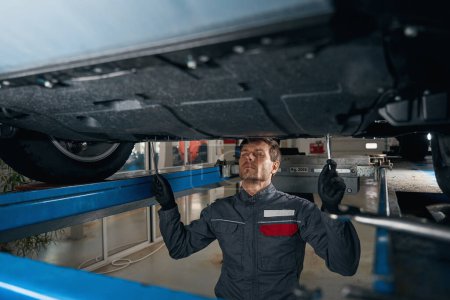 Photo for Mechanic in protective clothes standing in tire fitting under car and checking condition of the wheels - Royalty Free Image