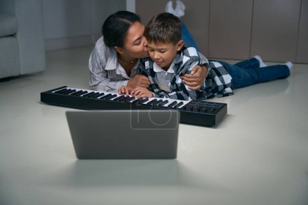 Téléchargez les photos : Loving african american mom kissing her teen son, they are having fun with digital piano and laptop - en image libre de droit