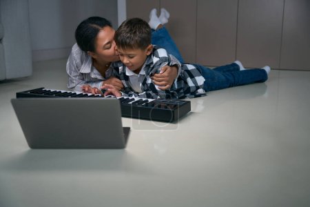 Photo for African american mother hugging and kissing her teenage son, they sit comfortably on the floor with digital piano and laptop - Royalty Free Image