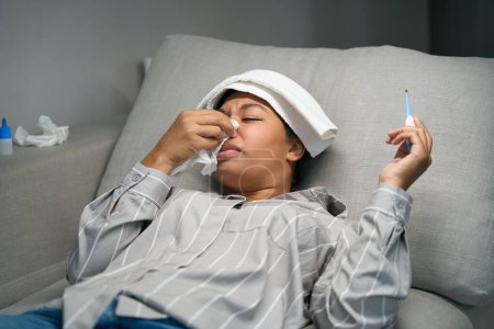Téléchargez les photos : Femme afro-américaine couchée sur le canapé avec un nez qui coule et une forte fièvre, elle a un thermomètre et des médicaments - en image libre de droit