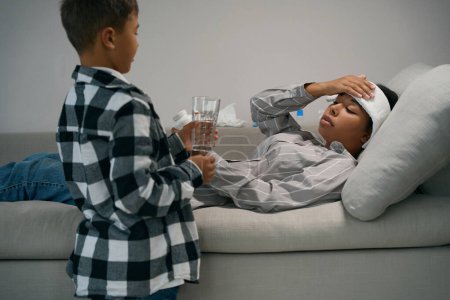 Téléchargez les photos : Garçon a apporté un verre d'eau à sa mère malade, la femme s'allonge sur le canapé avec un mal de tête - en image libre de droit