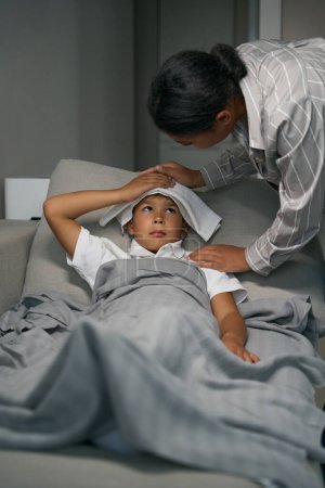 Foto de Adolescente sufre de un dolor de cabeza, se acuesta en el sofá, su madre pone una compresa en la frente - Imagen libre de derechos