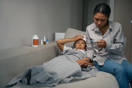 Foto de Young woman sits on a sofa with a thermometer next to a sick child, they use a compress and medicine - Imagen libre de derechos