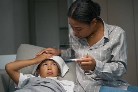 Photo for Kind mother takes care of a sick boy, she measures his temperature and makes a compress on his forehead - Royalty Free Image