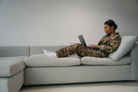 Photo for Young soldier woman is typing something in a laptop, she is comfortably seated on the couch - Royalty Free Image