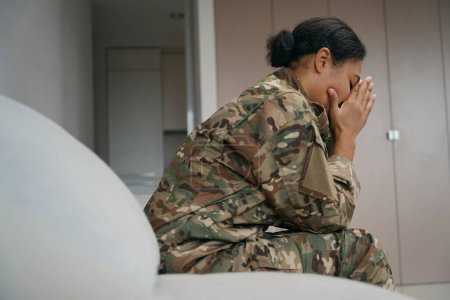 Foto de Tired african american female soldier sits alone in a bright room, she is wearing camouflage clothes - Imagen libre de derechos