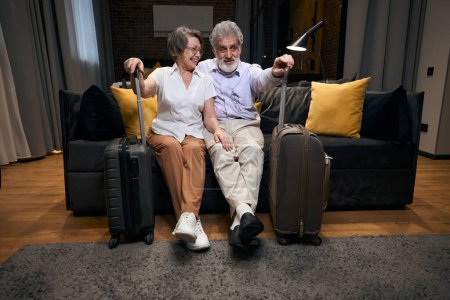 Photo for Elderly female and man sitting on the sofa in hotel, holding travel bag and talking - Royalty Free Image