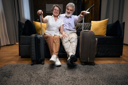 Photo for Elderly lady and man sitting on the sofa in hotel, holding travel bag and looking at the camera - Royalty Free Image