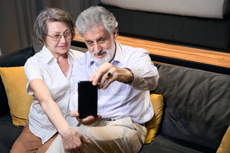 Téléchargez les photos : Vieille dame et homme assis sur le canapé de l'hôtel, mâle tenant téléphone portable et prendre des photos - en image libre de droit