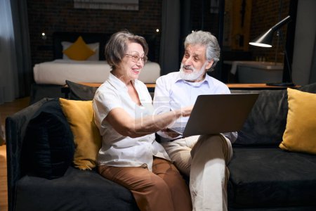 Foto de Happy elderly man and woman sitting on sofa in hotel, looking at laptop and doing online shopping - Imagen libre de derechos