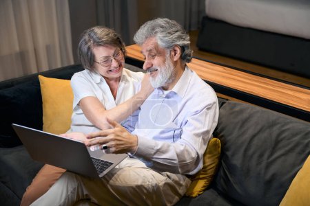 Photo for Elderly man and woman sitting on sofa in hotel, looking at laptop, talking and doing online shopping - Royalty Free Image