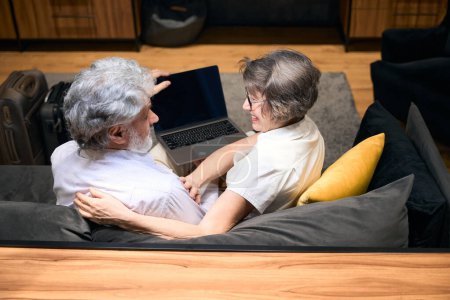 Foto de Hombre y mujer mayores sentados en el sofá del hotel, sosteniendo el portátil, mirándose - Imagen libre de derechos