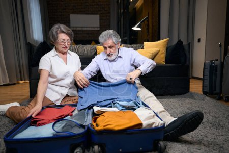 Téléchargez les photos : Elderly male and woman sitting on floor in hotel, man putting clothes in suitcase - en image libre de droit