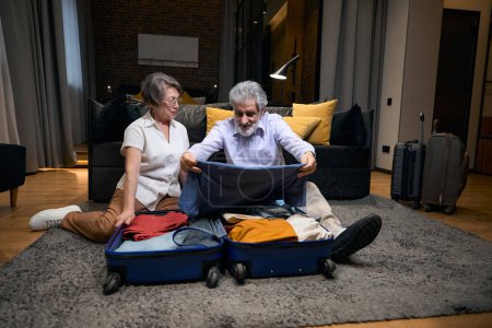 Téléchargez les photos : Elderly man and woman sitting on floor in hotel, male putting clothes in suitcase - en image libre de droit