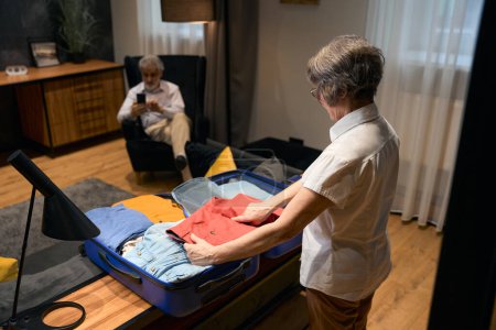 Photo for Elderly woman standing in hotel, putting clothes in suitcase, male sitting and looking at phone - Royalty Free Image