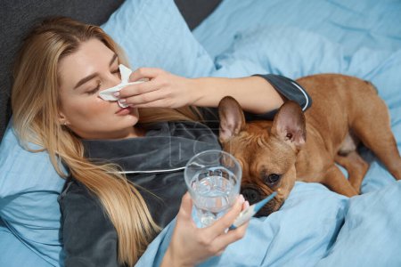 Téléchargez les photos : Malade femme tenant thermomètre numérique et verre d'eau dans les mains tout en soufflant du nez dans le mouchoir - en image libre de droit