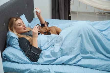 Téléchargez les photos : Femme malade couchée au lit et regardant le thermomètre numérique en présence de son bouledogue français - en image libre de droit