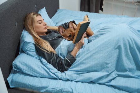 Photo for Young female lying in bed with pet and open book in hands - Royalty Free Image