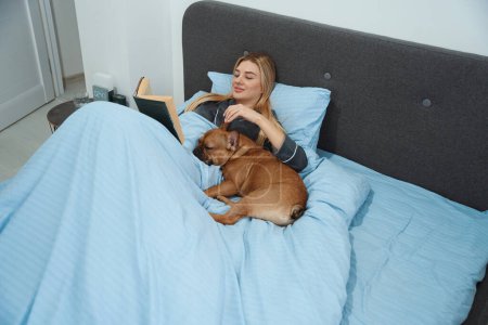 Photo for Smiling pleased young female stroking her pet and reading novel in bed - Royalty Free Image