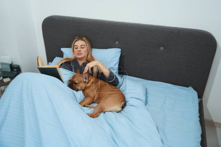 Photo for Calm focused young woman stroking her French bulldog while reading novel in bedroom - Royalty Free Image