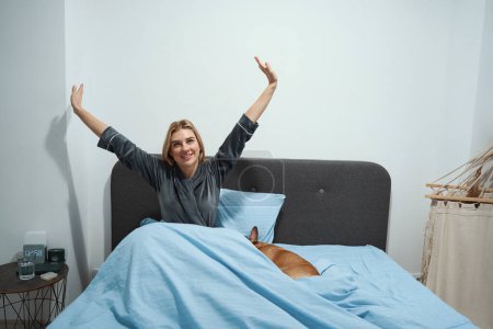 Photo for Smiling young woman wrapped in duvet stretching in cozy bed while her dog sleeping - Royalty Free Image