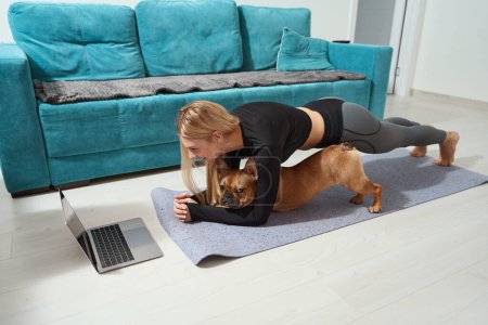 Téléchargez les photos : Femme sportive souriante et son chien faisant planche avant-bras avant ordinateur portable sur tapis de yoga - en image libre de droit