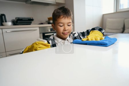 Téléchargez les photos : Enfant dans des gants de protection aide au nettoyage de la maison, il lave la surface de la cuisine - en image libre de droit