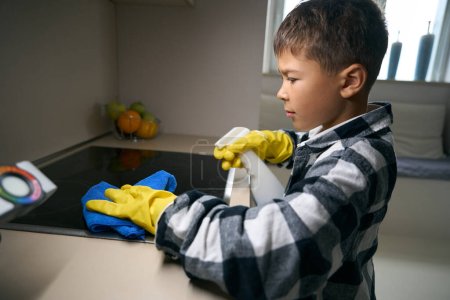 Teenager in Schutzhandschuhen wäscht vorsichtig den Küchenherd, er benutzt einen Lappen und eine Sprühflasche