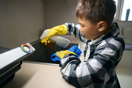 Téléchargez les photos : Teenager gently sprays detergent on a kitchen stove, he uses a rag and a spray bottle - en image libre de droit