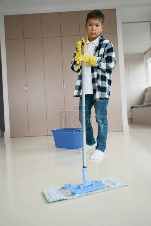 Téléchargez les photos : Neat boy washes the floor in the living room with a mop, a teenager in clean casual clothes - en image libre de droit