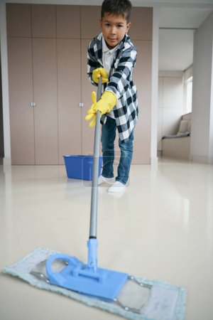 Foto de Lindo niño fregando el suelo en la sala de estar con una fregona, niño en ropa casual limpia - Imagen libre de derechos