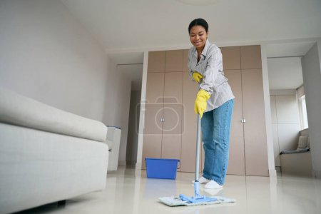 Téléchargez les photos : Smiling emale african american mopping the floor in the living room, woman in comfortable casual clothes - en image libre de droit