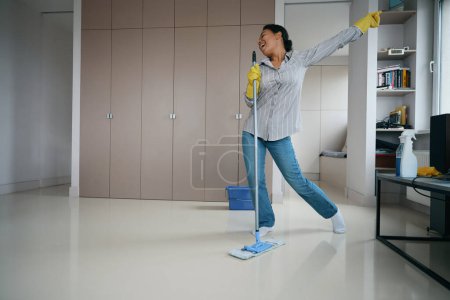 Téléchargez les photos : Optimistic young woman started a cheerful cleaning in the living room, she washes the floor, sings and dances - en image libre de droit