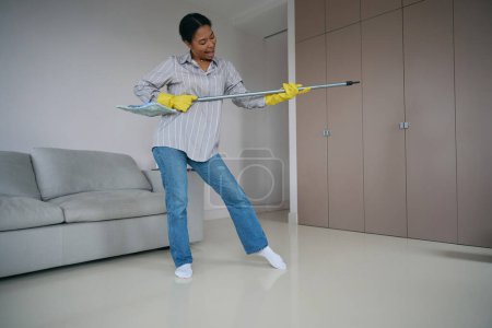 Téléchargez les photos : Joyeux jeune femme a commencé un nettoyage amusant dans la maison, elle lave le sol, chante et joue de la serpillière - en image libre de droit