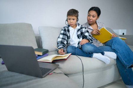 Photo for Mom helps a student at home in online learning, the boy uses a laptop, headset, books - Royalty Free Image