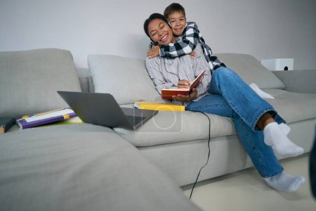 Foto de Niño abraza a su madre, que está estudiando cursos en línea en casa, la mujer utiliza los auriculares - Imagen libre de derechos