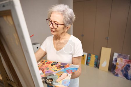 Foto de Sofisticada dama está pintando un cuadro en habitación luminosa, una mujer tiene una paleta y un cepillo en sus manos - Imagen libre de derechos