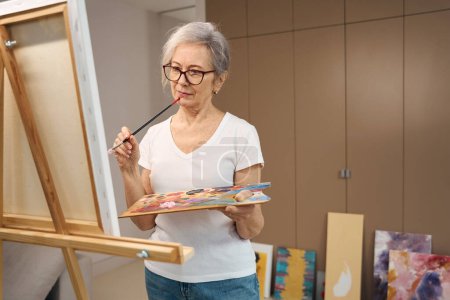 Téléchargez les photos : Slender elderly lady paints a picture in a spacious room, her previous works are by the closet - en image libre de droit