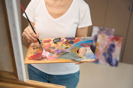 Foto de Mujer mayor sostiene una paleta y un pincel en sus manos, sus pinturas terminadas están cerca del armario - Imagen libre de derechos