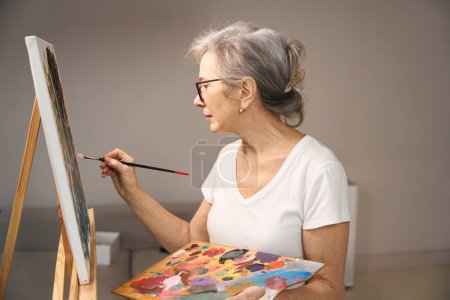 Foto de Dama de pelo gris en ropa casual se para frente a un caballete, ella pinta un cuadro - Imagen libre de derechos