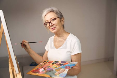 Foto de Mujer sofisticada de pelo gris en gafas se para frente a un caballete, ella pinta un cuadro - Imagen libre de derechos