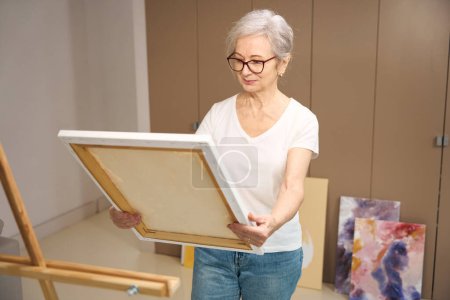 Foto de Una anciana sonriente sostiene un lienzo en una camilla en sus manos, pinturas terminadas junto al armario - Imagen libre de derechos