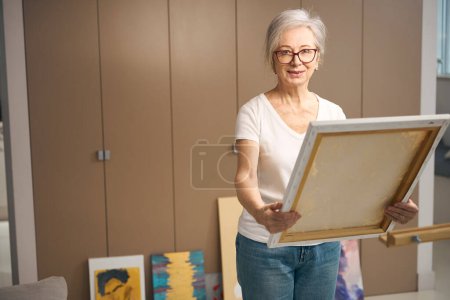 Photo for Happy elderly woman holds a canvas on a stretcher in her hands, her finished paintings are near the closet - Royalty Free Image