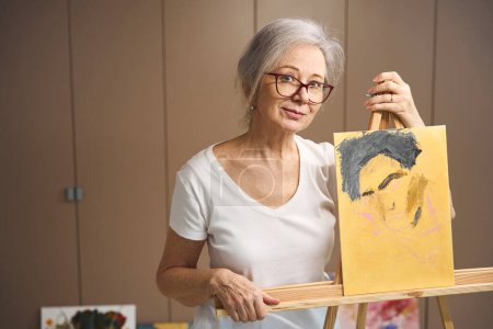 Téléchargez les photos : Elderly pensioner stands at an easel with an unfinished portrait, drawing her hobby - en image libre de droit